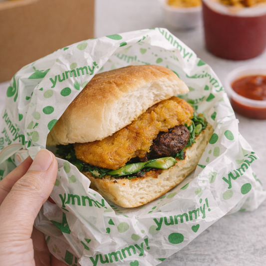 A hand holding a sandwich wrapped in white paper printed with the green 'yummy!' logo, with blurred food containers in the background.