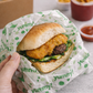 A hand holding a sandwich wrapped in white paper printed with the green 'yummy!' logo, with blurred food containers in the background.